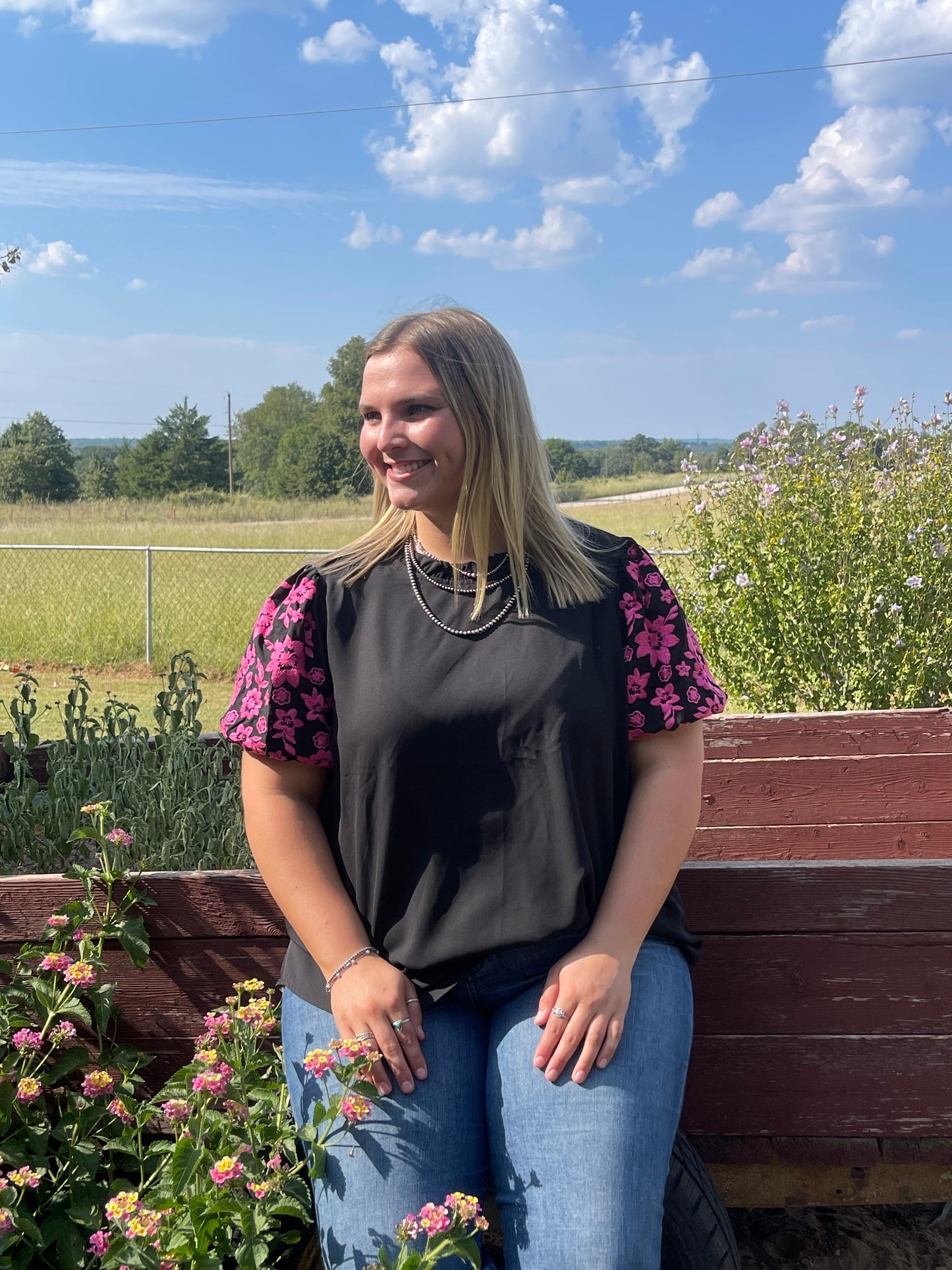 Woman wearing a black short puff sleeve shirt with pink embroidered flowers on the sleeves, in a size 1X