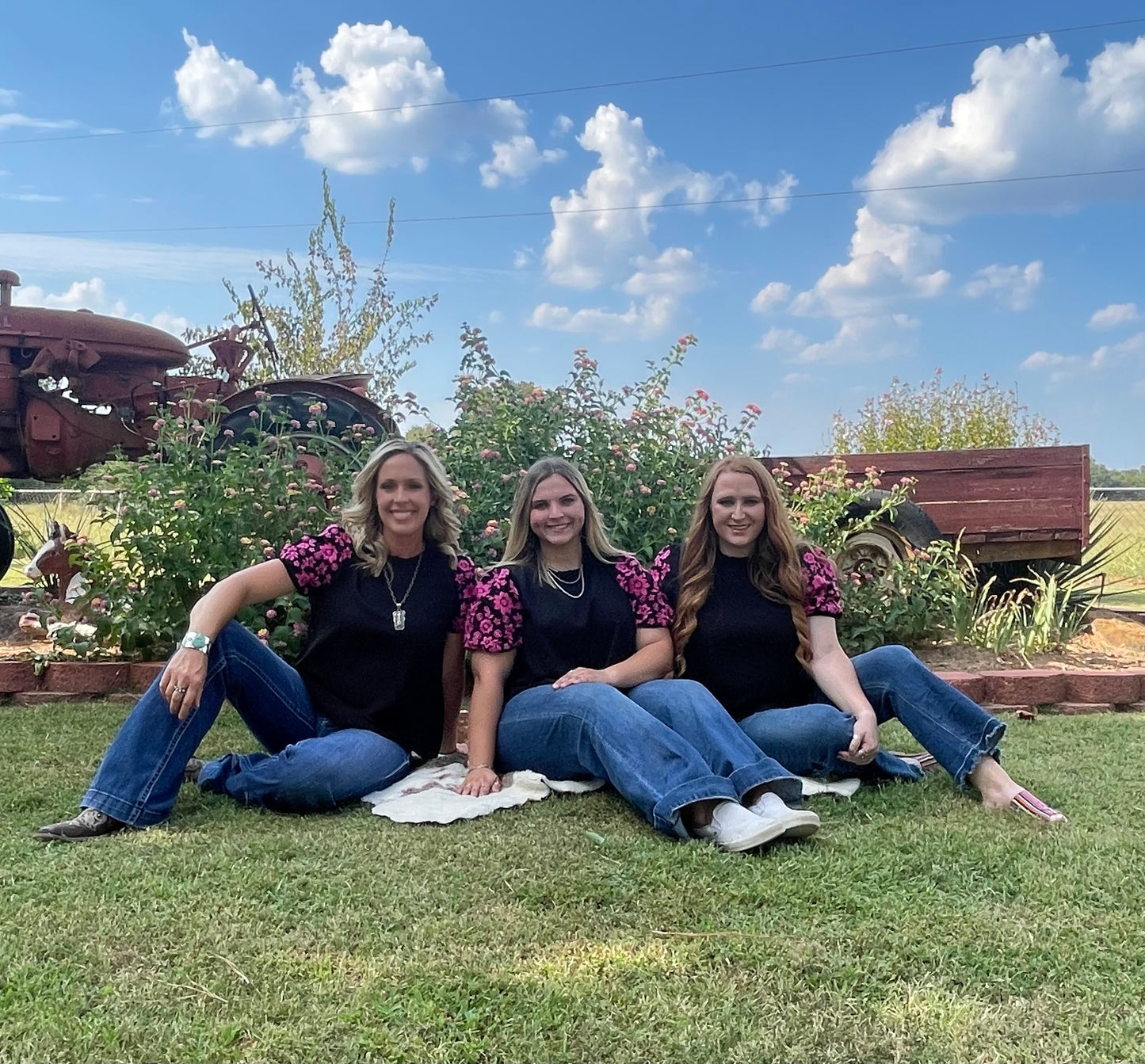 Three women wearing a black short puff sleeve shirt with pink embroidered flowers on the sleeves, in three different sizes