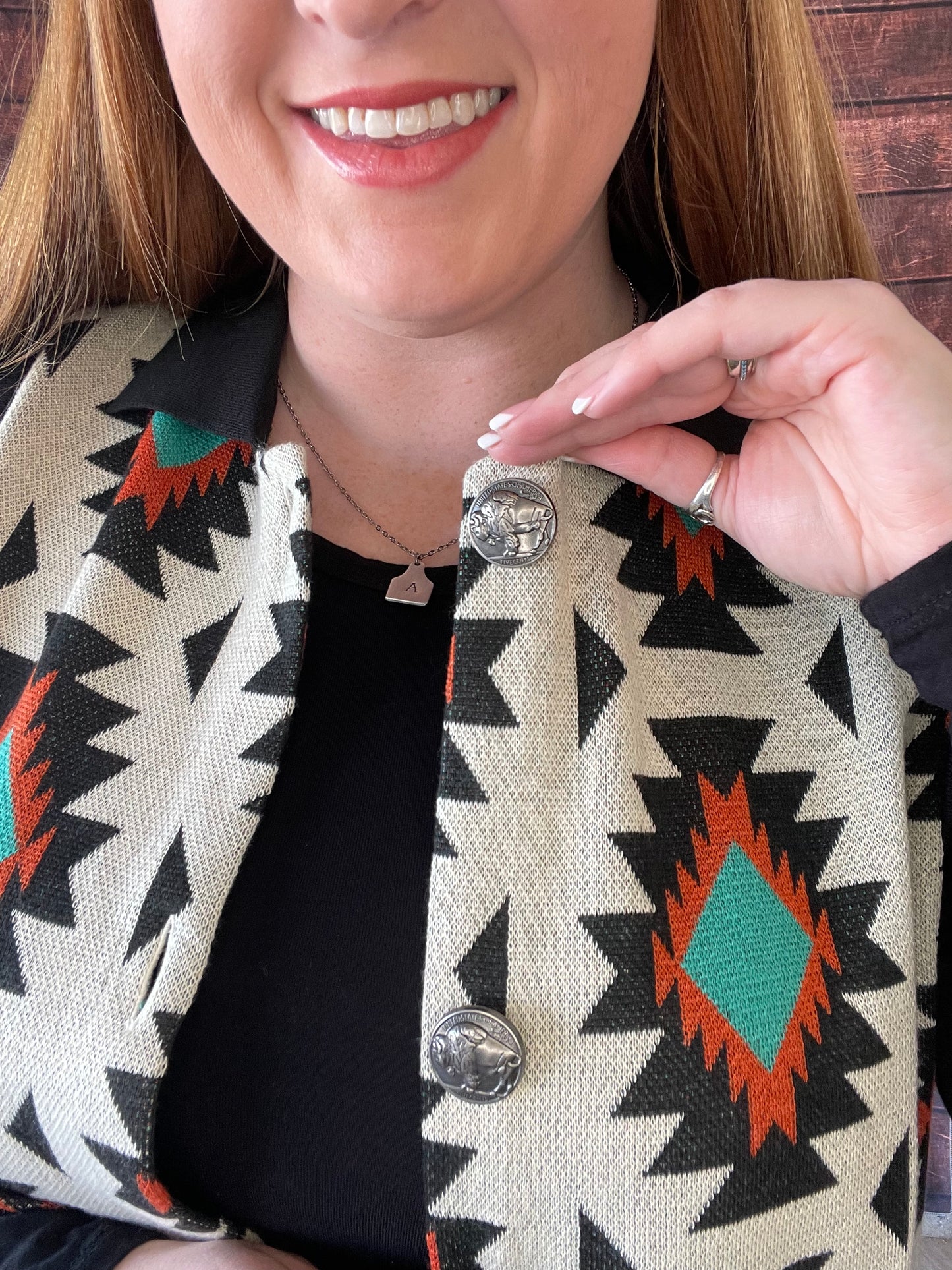 Close up of model wearing cream sweater vest with rust and turquoise Aztec detailing along with black collar and buffalo nickel buttons in size Large showing button detailing