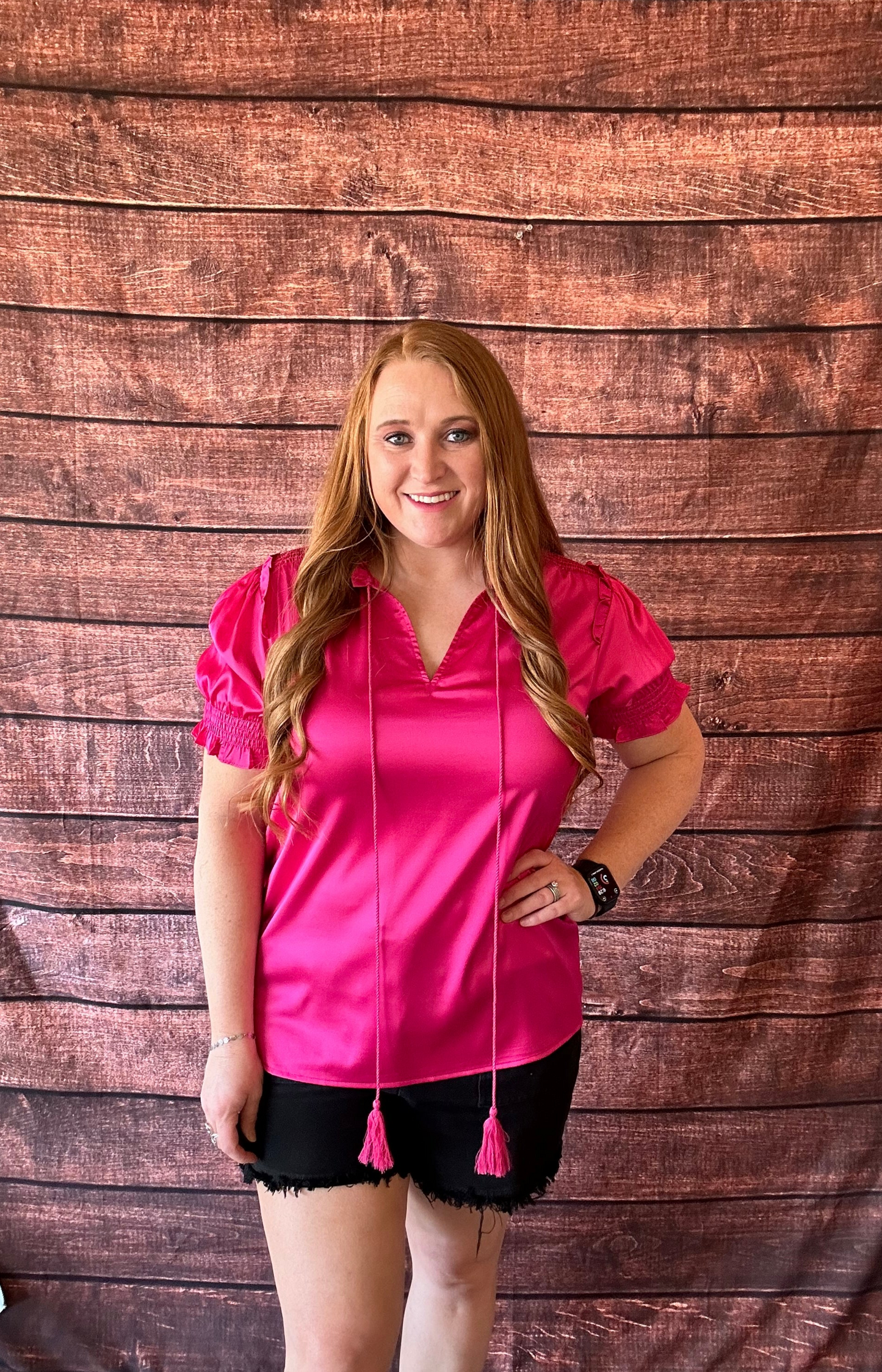 Model wearing bright pink short sleeve blouse with a shiny satin like finish with black shorts