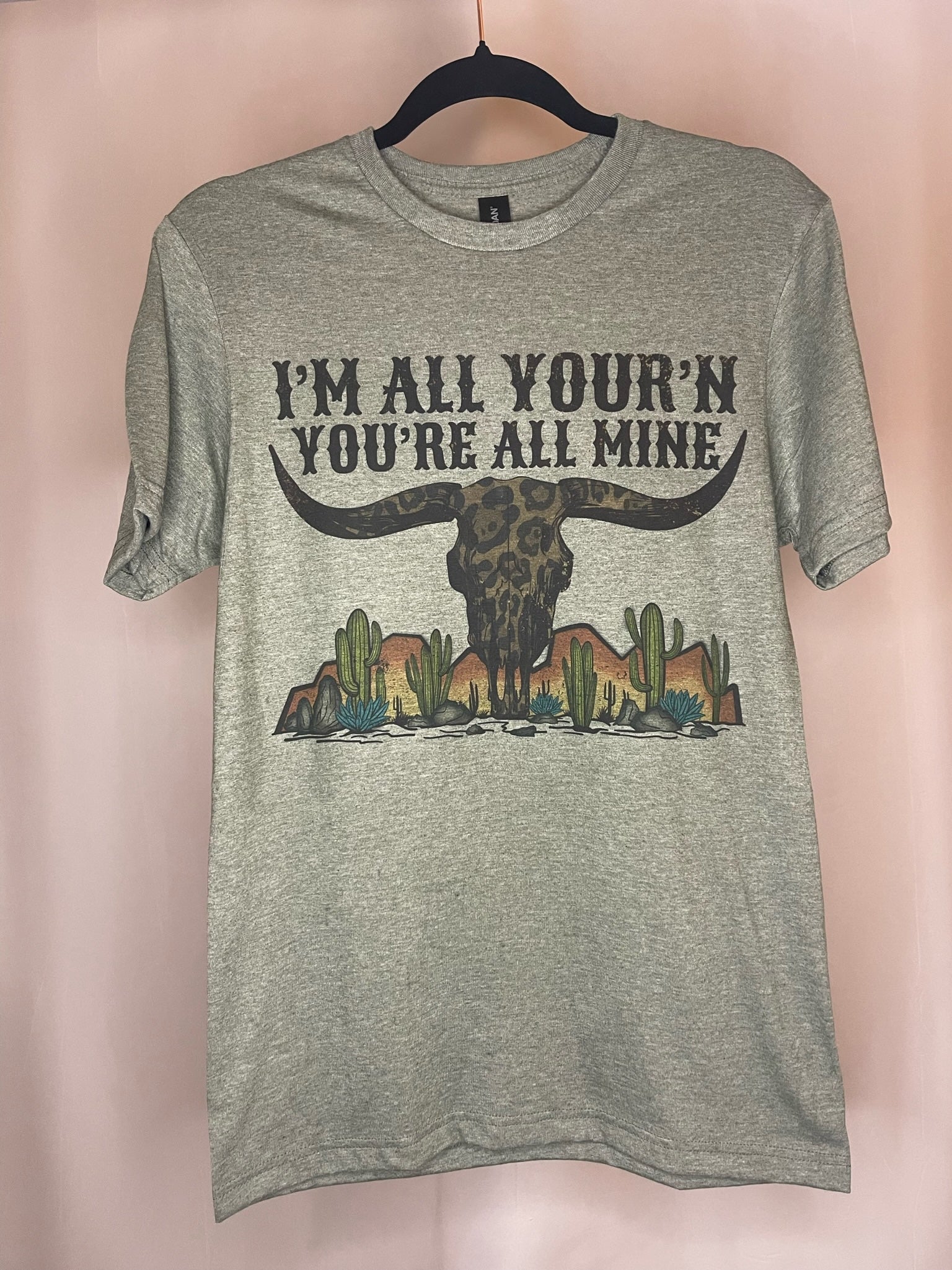 Heather Olive t-shirt with desert mountains and cactus scene, a cheetah cow skull with horns, and the words "I'm all your'n you're all mine" 