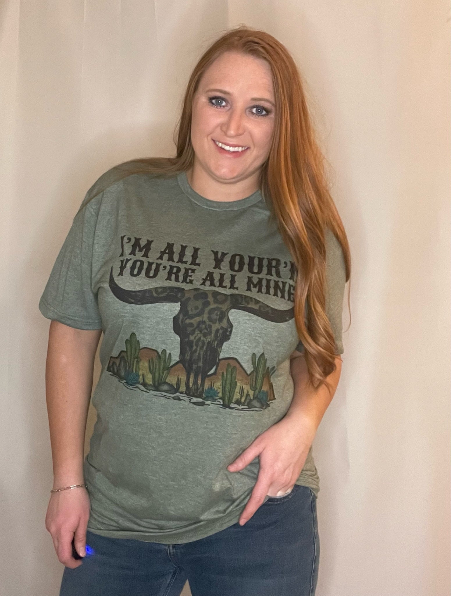 Woman wearing heather olive t-shirt with desert mountains and cactus scene, a cheetah cow skull with horns, and the words "I'm all your'n you're all mine"